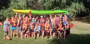 Grupa kajakarzy w kamizelkach ratunkowych z wiosłami w rękach stoją trzymając w górze kajak.